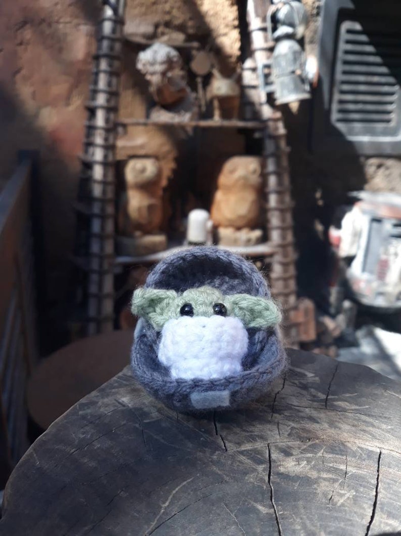 May include: A small green crocheted character with large eyes, wearing a gray crocheted hooded robe, sitting on a wooden surface.