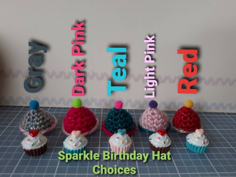 May include: Five crocheted cupcake hats in different colors: gray, dark pink, teal, light pink, and red. Each hat has a small pom-pom on top and a cupcake charm. The text "Sparkle Birthday Hat Choices" is below the hats.