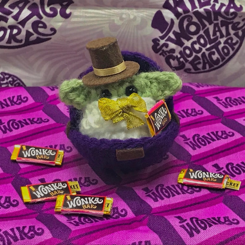 May include: A handmade, crocheted character wearing a brown top hat and a gold bow tie, holding a miniature Wonka Bar. The character is seated in a purple container, surrounded by miniature Wonka Bars on a purple Wonka-themed fabric.