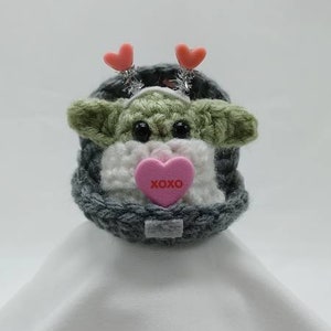 Puede incluir: Un juguete de peluche de crochet verde y blanco con un gorro de punto gris y un coraz&oacute;n rosa con el texto "XOXO". El juguete tiene dos corazones rosas en la cabeza.