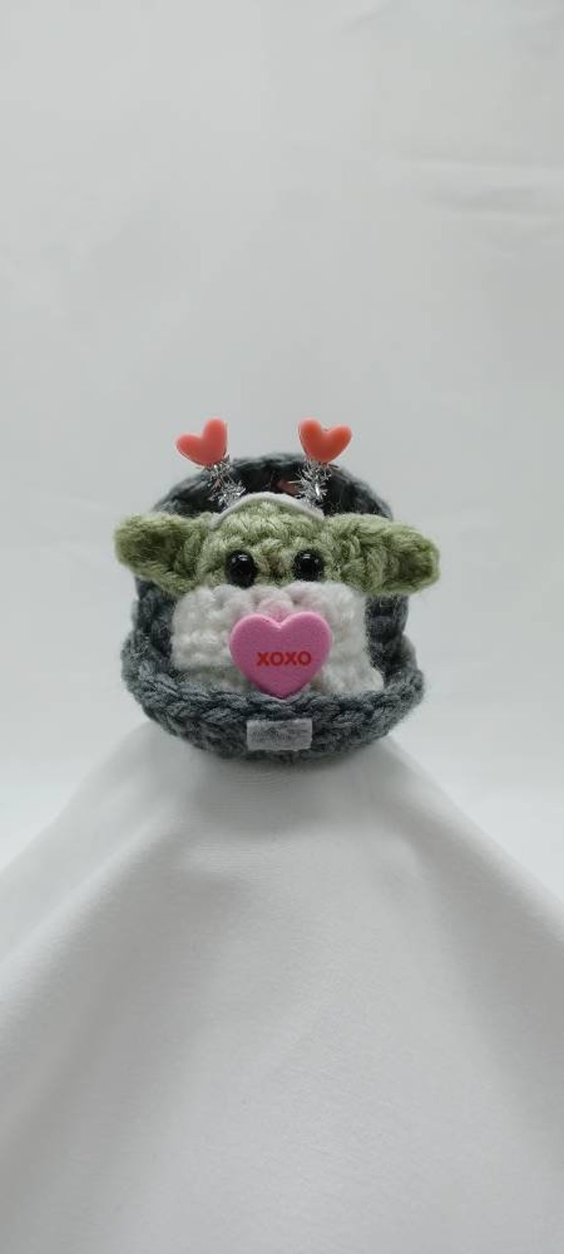 Puede incluir: Un juguete de peluche de crochet verde y blanco con un gorro de punto gris y un coraz&oacute;n rosa con el texto "XOXO". El juguete tiene dos corazones rosas en la cabeza.