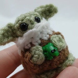 Peut inclure: Une petite cr&eacute;ature crochet&eacute;e avec une peau en laine vert clair, de grands yeux en plastique noir et des oreilles pro&eacute;minentes. Elle porte un v&ecirc;tement en crochet marron avec un col blanc et tient une minuscule figurine de grenouille verte.