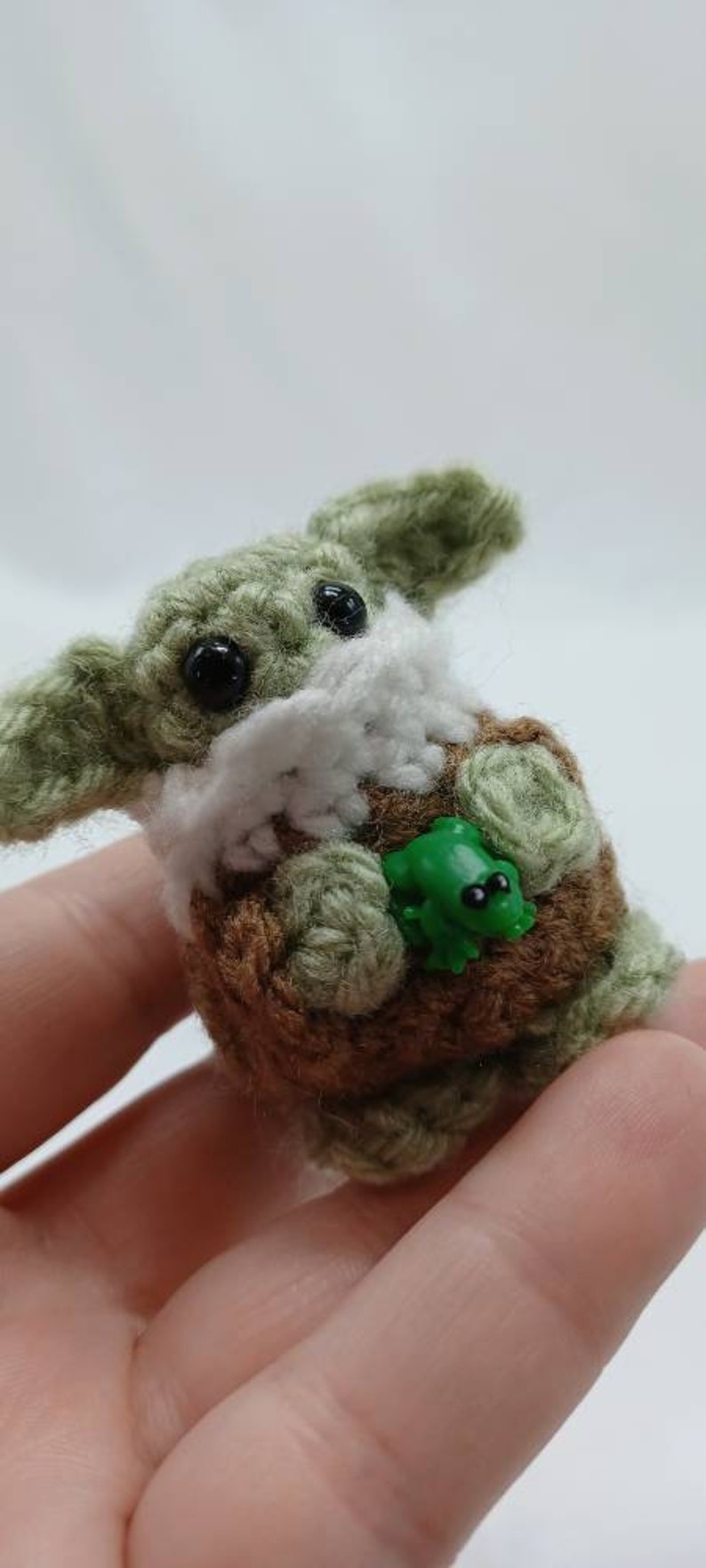 Peut inclure: Une petite cr&eacute;ature crochet&eacute;e avec une peau en laine vert clair, de grands yeux en plastique noir et des oreilles pro&eacute;minentes. Elle porte un v&ecirc;tement en crochet marron avec un col blanc et tient une minuscule figurine de grenouille verte.