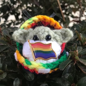 May include: A crocheted green and white character wearing a rainbow flag pin and a multi-colored crocheted scarf.