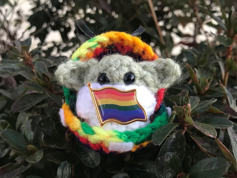May include: A crocheted green and white character wearing a rainbow flag pin and a multi-colored crocheted scarf.