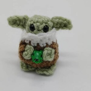 Peut inclure: Une petite cr&eacute;ature crochet&eacute;e avec une peau en laine vert clair, de grands yeux en plastique noir et de larges oreilles. Elle porte un v&ecirc;tement textur&eacute; marron avec un col blanc et tient une minuscule figurine de grenouille en plastique vert.