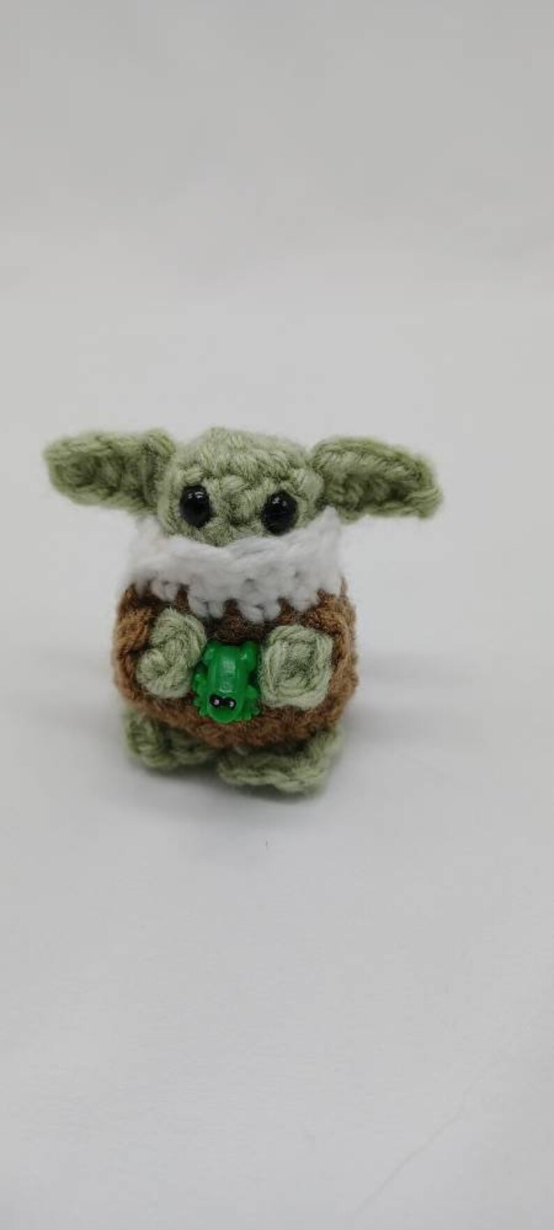 Peut inclure: Une petite cr&eacute;ature crochet&eacute;e avec une peau en laine vert clair, de grands yeux en plastique noir et de larges oreilles. Elle porte un v&ecirc;tement textur&eacute; marron avec un col blanc et tient une minuscule figurine de grenouille en plastique vert.