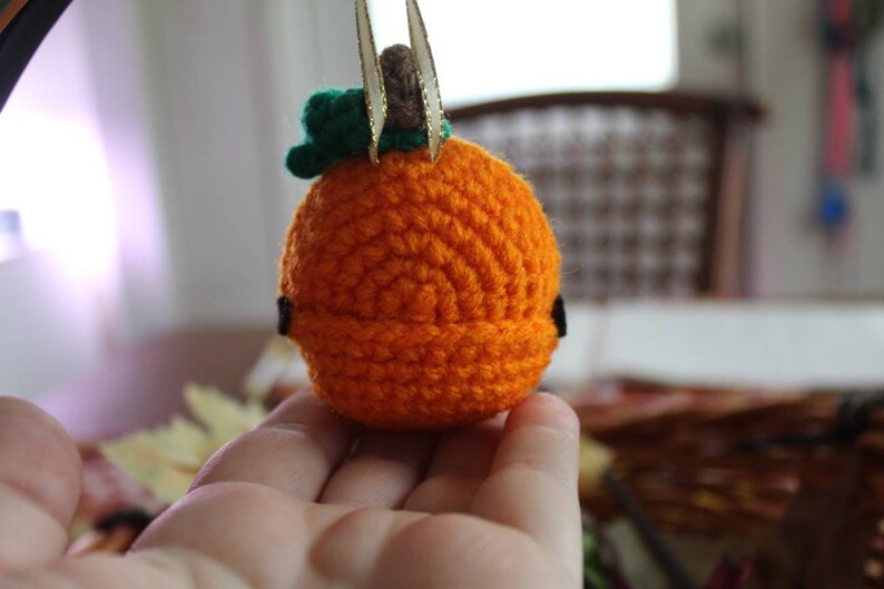 May include: A small crocheted pumpkin with a green stem and gold accents. The pumpkin is orange and has a textured surface.