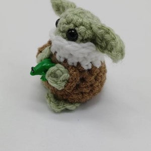 Peut inclure: Une petite figurine au crochet avec une peau vert clair, de grandes oreilles et des yeux en perles noires. Elle porte un col blanc et un corps textur&eacute; marron. La figurine tient une petite grenouille en plastique vert vif dans ses mains.
