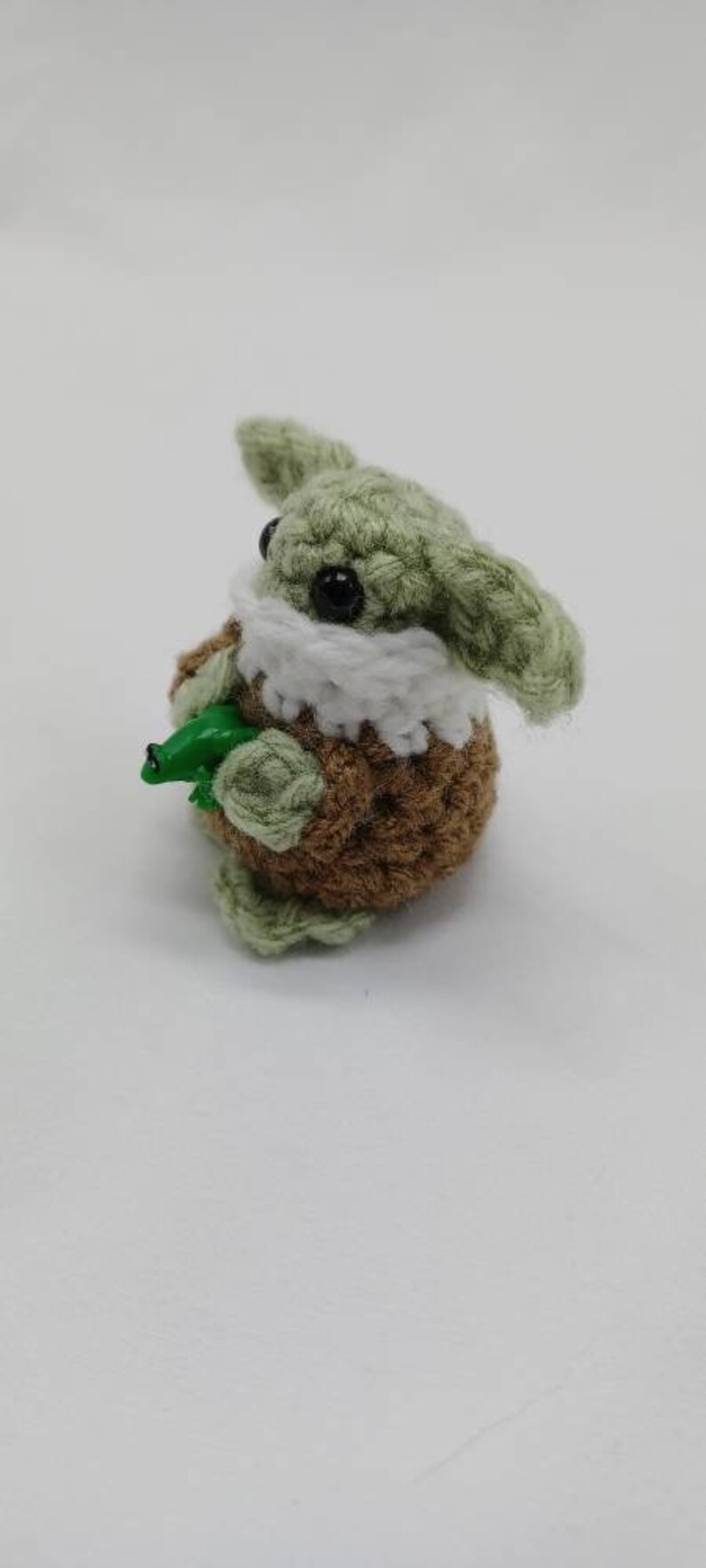 Peut inclure: Une petite figurine au crochet avec une peau vert clair, de grandes oreilles et des yeux en perles noires. Elle porte un col blanc et un corps textur&eacute; marron. La figurine tient une petite grenouille en plastique vert vif dans ses mains.
