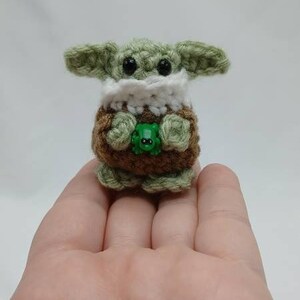 Peut inclure: Une petite figurine crochet&eacute;e &agrave; la main avec une t&ecirc;te vert clair, de grands yeux noirs, de larges oreilles et un col blanc. Le corps est marron avec des pieds et des mains vert clair, tenant une minuscule grenouille en plastique vert vif.