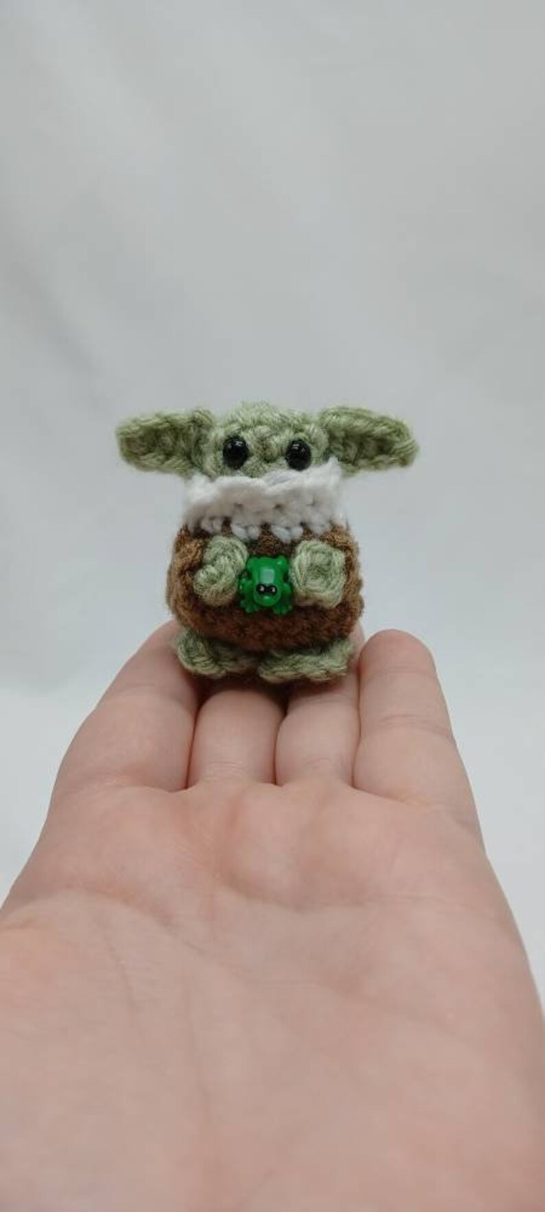 Peut inclure: Une petite figurine crochet&eacute;e &agrave; la main avec une t&ecirc;te vert clair, de grands yeux noirs, de larges oreilles et un col blanc. Le corps est marron avec des pieds et des mains vert clair, tenant une minuscule grenouille en plastique vert vif.