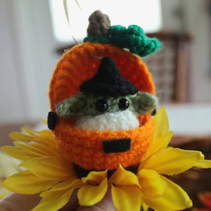 May include: A small crocheted green and white character wearing a black witch hat sits inside a crocheted orange pumpkin. The pumpkin is sitting on a yellow sunflower.