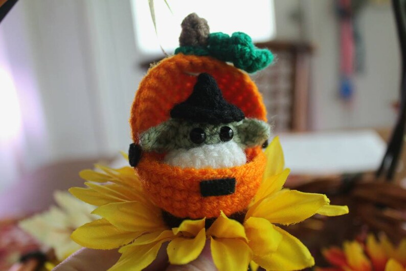 May include: A small crocheted green and white character wearing a black witch hat sits inside a crocheted orange pumpkin. The pumpkin is sitting on a yellow sunflower.