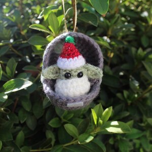 May include: A crocheted Christmas ornament featuring a green and white character wearing a red and white Santa hat. The character is inside a gray crocheted pod.