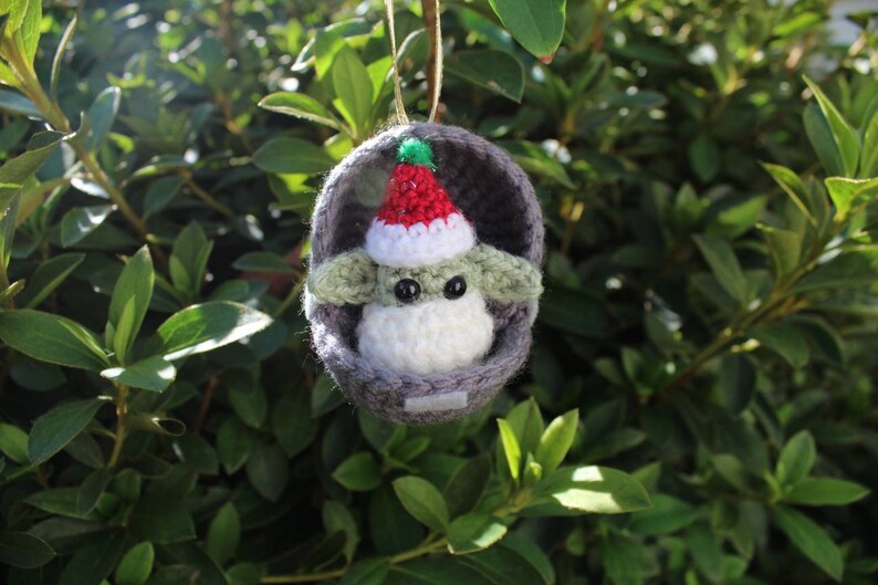 May include: A crocheted Christmas ornament featuring a green and white character wearing a red and white Santa hat. The character is inside a gray crocheted pod.
