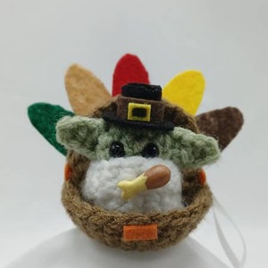 Puede incluir: Un juguete de peluche tejido a crochet con forma de beb&eacute; Yoda que lleva un sombrero de peregrino y sostiene una pierna de pavo. El juguete est&aacute; decorado con un tocado de plumas de pavo de color marr&oacute;n, verde, amarillo y rojo.