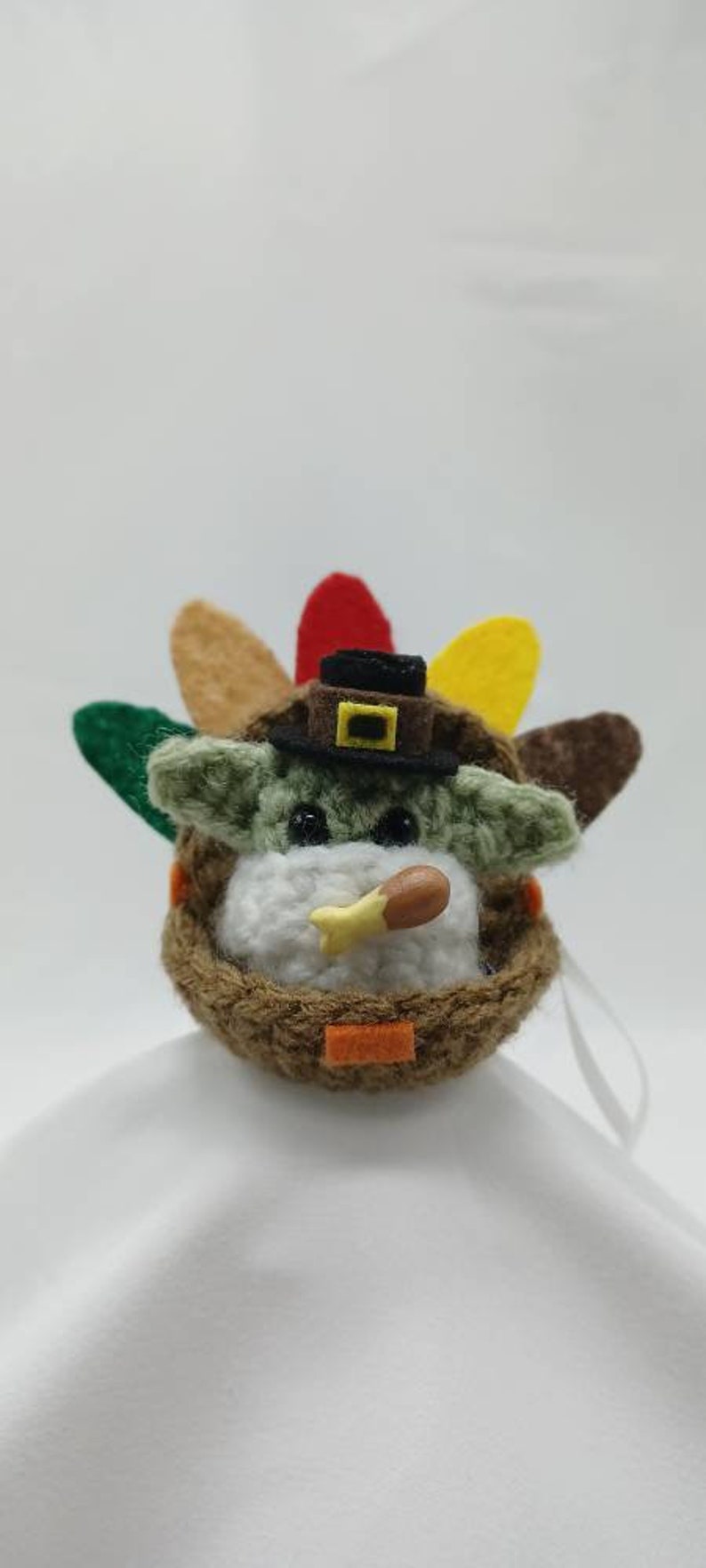Puede incluir: Un juguete de peluche tejido a crochet con forma de beb&eacute; Yoda que lleva un sombrero de peregrino y sostiene una pierna de pavo. El juguete est&aacute; decorado con un tocado de plumas de pavo de color marr&oacute;n, verde, amarillo y rojo.