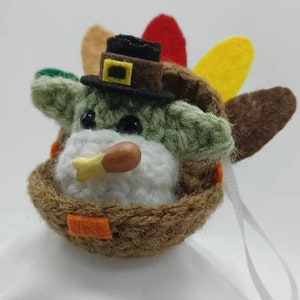 Puede incluir: Un adorno de pavo de ganchillo con un sombrero marr&oacute;n y una cara blanca. El pavo tiene un cuerpo marr&oacute;n con plumas verdes y amarillas. El adorno tiene una cuerda blanca para colgar.