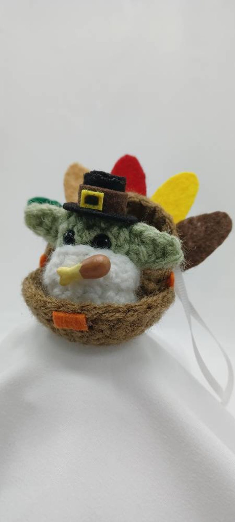 Puede incluir: Un adorno de pavo de ganchillo con un sombrero marr&oacute;n y una cara blanca. El pavo tiene un cuerpo marr&oacute;n con plumas verdes y amarillas. El adorno tiene una cuerda blanca para colgar.