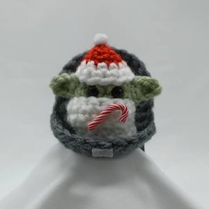 May include: A crocheted plush toy of a popular character wearing a red and white Santa hat and holding a candy cane. The toy is sitting on a white surface.