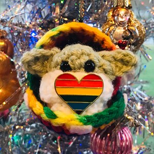 May include: A crocheted ornament shaped like a small creature with a rainbow heart pin on its chest. The ornament is hanging on a silver Christmas tree.