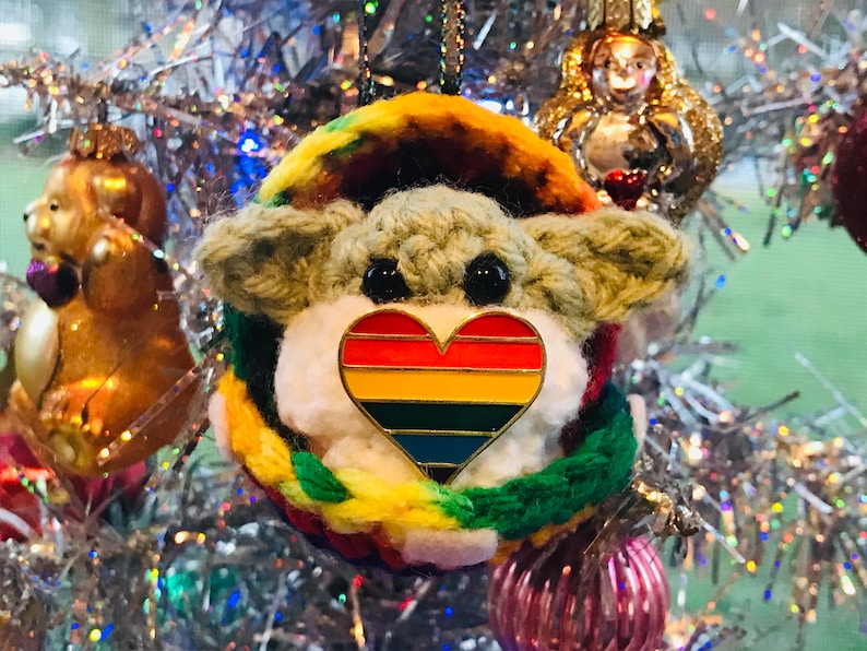 May include: A crocheted ornament shaped like a small creature with a rainbow heart pin on its chest. The ornament is hanging on a silver Christmas tree.