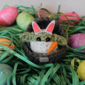 May include: A green crocheted Baby Yoda wearing pink bunny ears and holding a carrot, sitting in a gray crocheted basket, surrounded by Easter eggs in a bed of green grass.