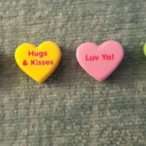 Puede incluir: Siete caramelos en forma de coraz&oacute;n con diferentes mensajes. Los caramelos son blancos, naranjas, amarillos, rosas, verdes y rosa claro. Los mensajes en los caramelos son "Love", "Cutie Pie", "Hugs & Kisses", "Luv Ya!", "Be Mine", "XOXO".