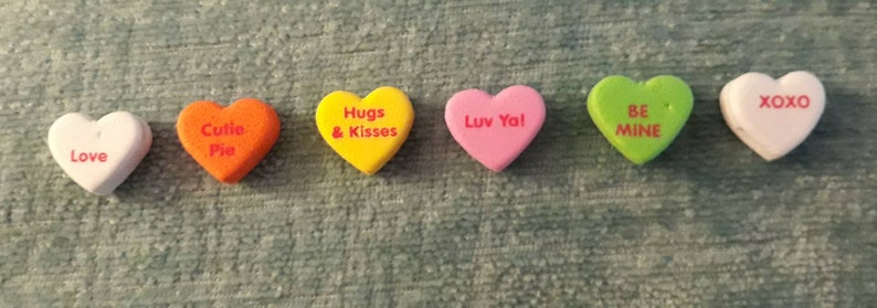 Puede incluir: Siete caramelos en forma de coraz&oacute;n con diferentes mensajes. Los caramelos son blancos, naranjas, amarillos, rosas, verdes y rosa claro. Los mensajes en los caramelos son "Love", "Cutie Pie", "Hugs & Kisses", "Luv Ya!", "Be Mine", "XOXO".
