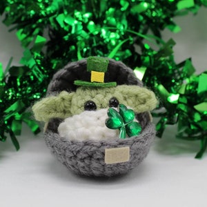 May include: A crocheted Yoda doll wearing a green hat with a gold buckle, holding a green shamrock. The doll is sitting in a grey crocheted egg-shaped basket.