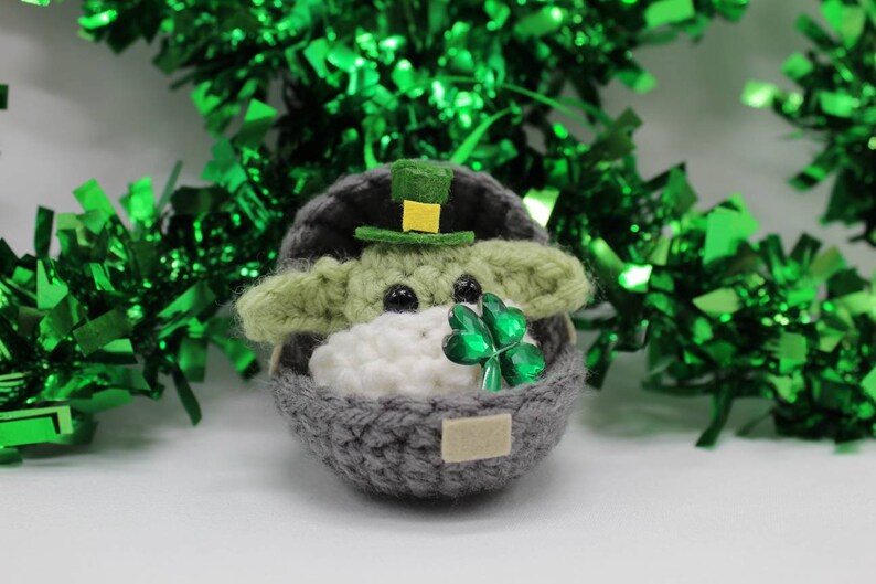 May include: A crocheted Yoda doll wearing a green hat with a gold buckle, holding a green shamrock. The doll is sitting in a grey crocheted egg-shaped basket.