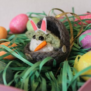 May include: A green crocheted character wearing a pink bunny ear headband and holding a carrot, sitting in a gray crocheted nest. The character is surrounded by colorful Easter eggs and green grass.