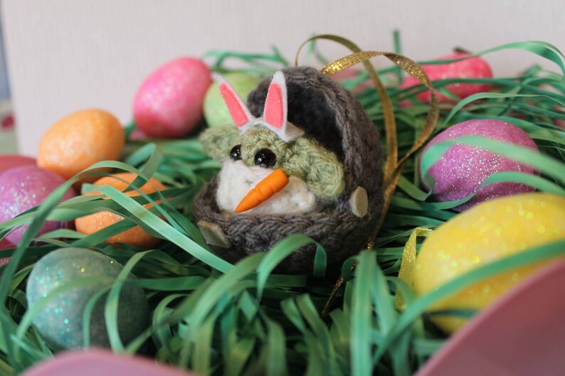 May include: A green crocheted character wearing a pink bunny ear headband and holding a carrot, sitting in a gray crocheted nest. The character is surrounded by colorful Easter eggs and green grass.