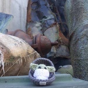 May include: A small green knitted toy character, resembling a baby, is sitting in a gray knitted cradle. The toy is placed on a green surface with a blurry background.