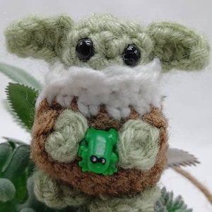 Peut inclure: Une petite cr&eacute;ature crochet&eacute;e &agrave; la peau vert clair, aux grands yeux noirs et aux oreilles larges. Elle porte un col blanc sur un corps textur&eacute; marron et tient une petite figurine de grenouille en plastique vert vif entre ses mains.