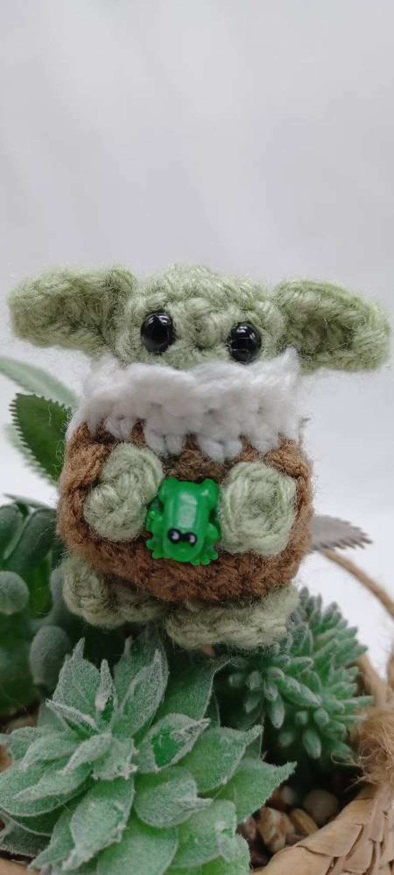 Peut inclure: Une petite cr&eacute;ature crochet&eacute;e &agrave; la peau vert clair, aux grands yeux noirs et aux oreilles larges. Elle porte un col blanc sur un corps textur&eacute; marron et tient une petite figurine de grenouille en plastique vert vif entre ses mains.