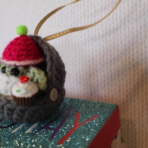 May include: A crocheted gray and green ornament of a creature wearing a pink hat and holding a cupcake. The ornament is hanging from a gold ribbon.