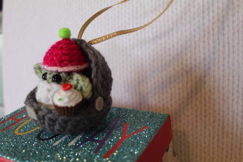 May include: A crocheted gray and green ornament of a creature wearing a pink hat and holding a cupcake. The ornament is hanging from a gold ribbon.