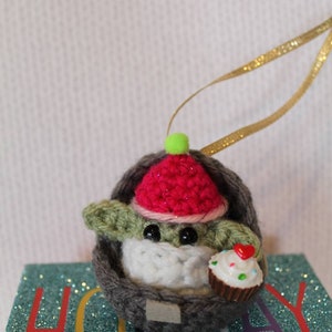May include: A crocheted Baby Yoda ornament wearing a red and pink hat with a green pom-pom. The ornament is holding a small cupcake with a red heart on top. The ornament is sitting on a blue and green glittery surface with the word "Holiday" printed on it.