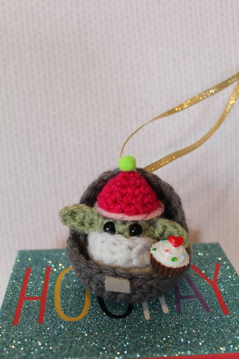 May include: A crocheted Baby Yoda ornament wearing a red and pink hat with a green pom-pom. The ornament is holding a small cupcake with a red heart on top. The ornament is sitting on a blue and green glittery surface with the word "Holiday" printed on it.