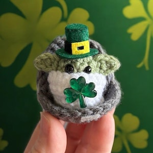 May include: A crocheted Yoda doll wearing a green leprechaun hat with a yellow band and holding a green shamrock. The doll is sitting in a hand against a green background with shamrock patterns.