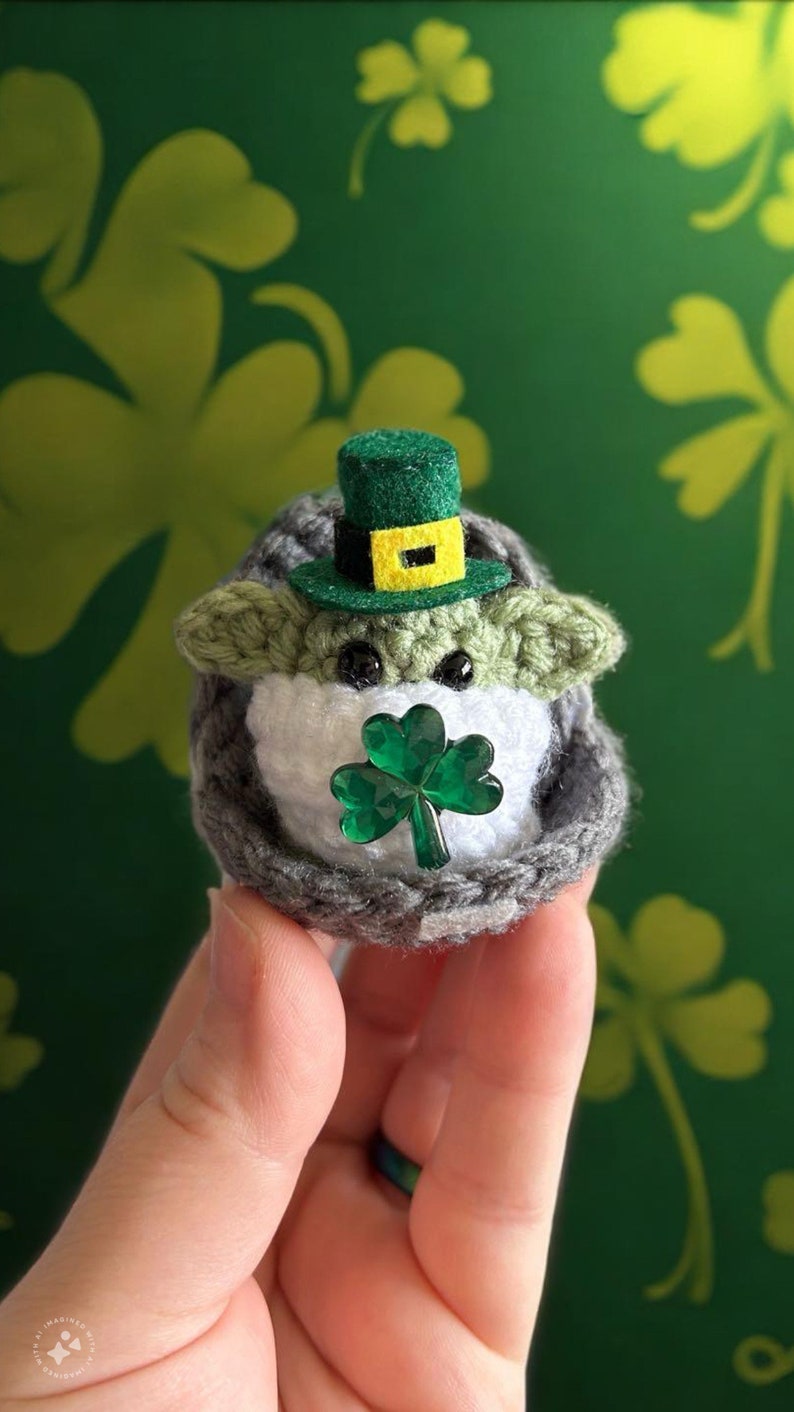 May include: A crocheted Yoda doll wearing a green leprechaun hat with a yellow band and holding a green shamrock. The doll is sitting in a hand against a green background with shamrock patterns.
