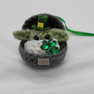May include: A crocheted Yoda ornament wearing a green hat with a yellow band and holding a green shamrock. The ornament is in a grey crocheted pot with a green ribbon.
