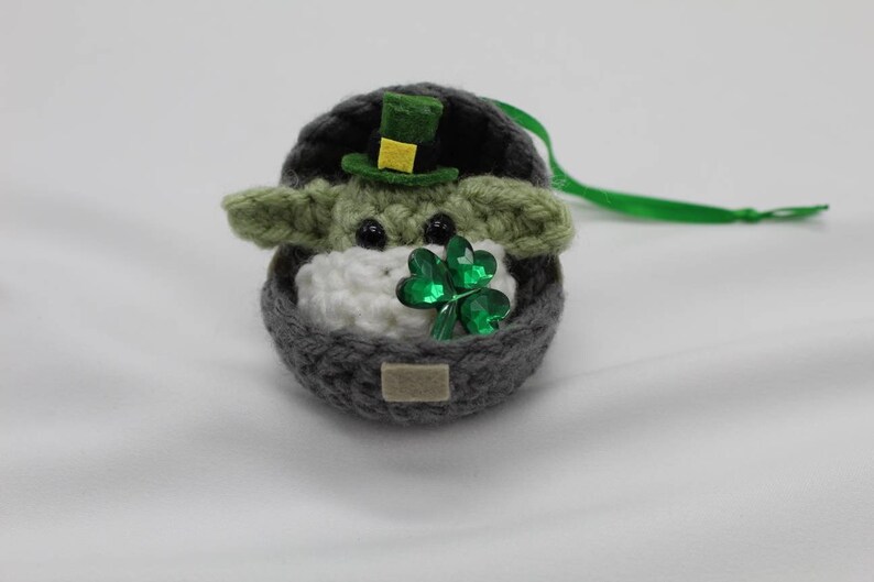 May include: A crocheted Yoda ornament wearing a green hat with a yellow band and holding a green shamrock. The ornament is in a grey crocheted pot with a green ribbon.