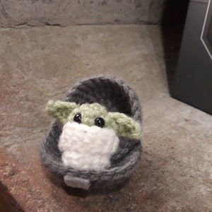 May include: A small crocheted plush toy of a green alien character, known as Baby Yoda, sitting in a gray crocheted pod.