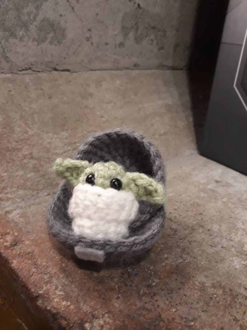 May include: A small crocheted plush toy of a green alien character, known as Baby Yoda, sitting in a gray crocheted pod.