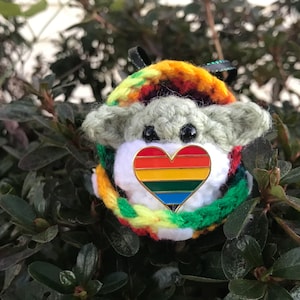 May include: A crocheted plush toy with a rainbow heart pin. The toy is green and white with a rainbow colored crocheted scarf.
