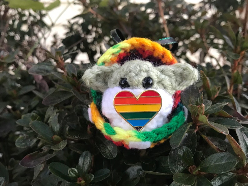 May include: A crocheted plush toy with a rainbow heart pin. The toy is green and white with a rainbow colored crocheted scarf.