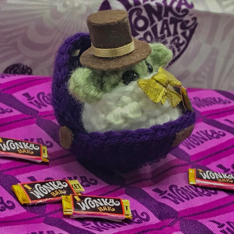 May include: A handmade, plush character with a green face, white body, and a purple outfit, wearing a brown top hat and a gold bow tie. Several miniature Wonka Bar candy wrappers are scattered around the character on a purple Wonka-themed fabric.